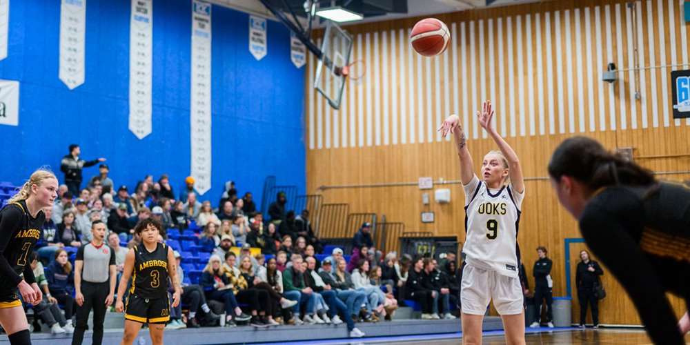 A basketball player in a white jersey is taking a free throw during a game, with opposing players in black jerseys and a crowd watching from the stands.