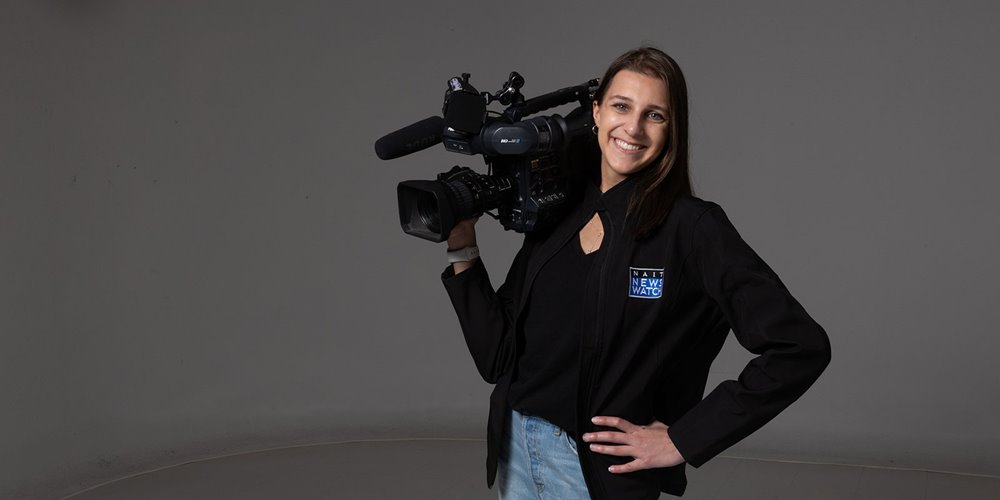 Person standing in a studio holding a professional video camera on their shoulder, wearing a black jacket with a NAIT News logo and light-colored jeans.