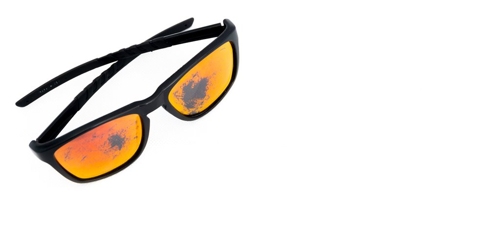 Black-framed sunglasses with orange-tinted lenses, showing dark smudges on the lenses, placed on a white background.