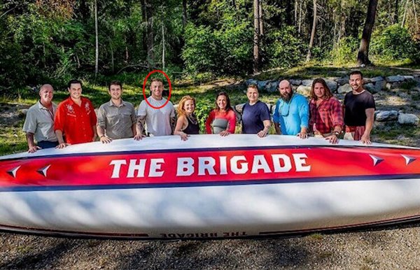 "Ten people standing behind a large canoe labeled 'THE BRIGADE' in a forested outdoor setting, with one individual circled in red."