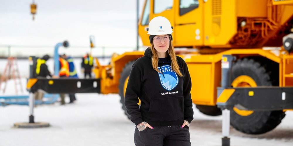 a woman in a white hard hat and dark blue hoodie with the words NAIT Crane and Hoist on it poses for a photo in front of a yellow crane.