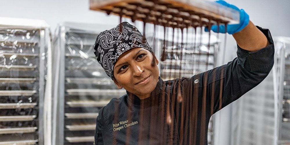 A person in a black chef's jacket labeled 'Priya Winsor, Corporate Chocolatier' and a patterned headscarf holds a tray with dripping chocolate. Racks covered with plastic sheets are visible in the background.