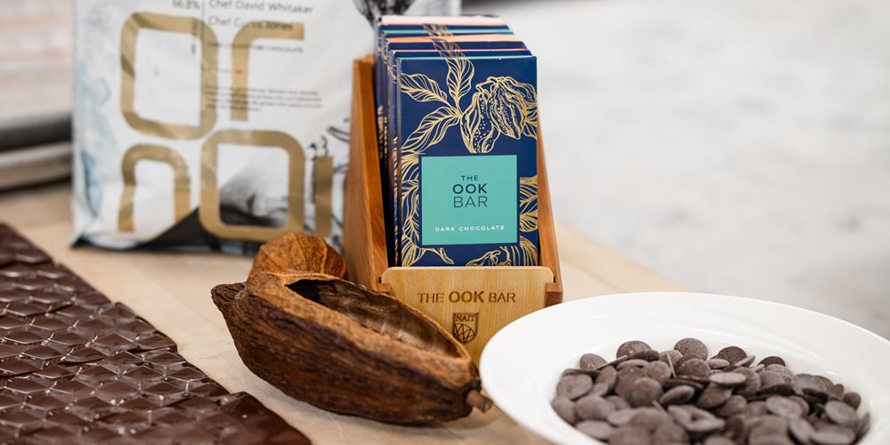 Dark chocolate bars in blue packaging displayed beside cacao pods, chocolate chips, and a chocolate slab on a light wooden surface.