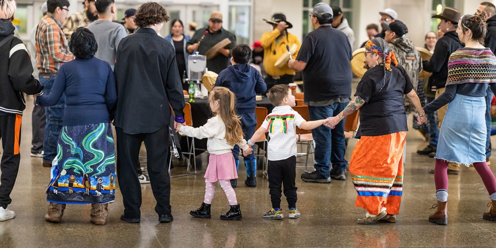 Children and adults holding hands in a circle inside a community hall, with people playing hand drums in the center and onlookers surrounding the group.