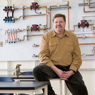 A person is sitting on a classroom table in front of a wall-mounted plumbing system display with copper pipes, valves, and gauges.