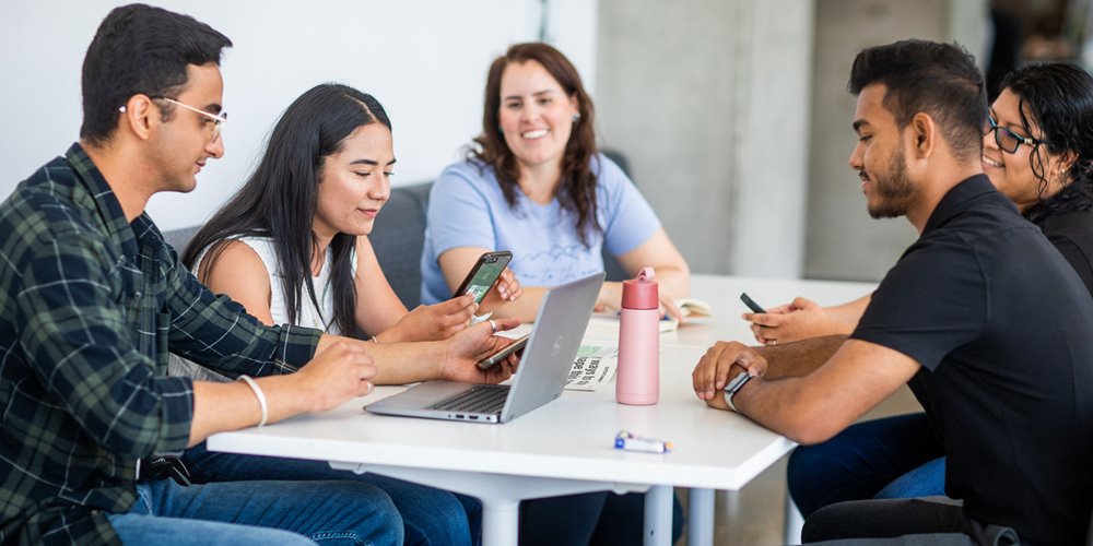 five students sit at a table talking and smiling. one woman is looking at a phone. a man has a laptop open