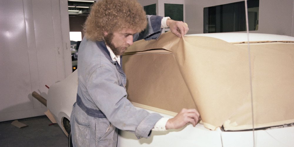Person wearing a work jacket applies masking paper to cover the side window of a white car inside an auto body shop, preparing the vehicle for painting.