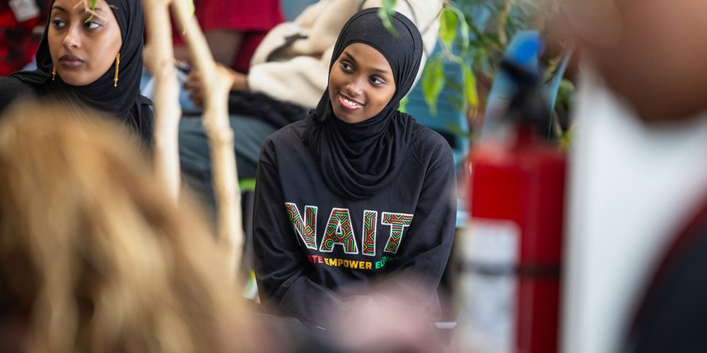 Person seated indoors wearing a black NAIT hoodie, surrounded by other people and campus furnishings in a casual gathering space.