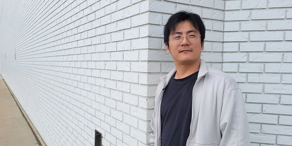 a man in a white jacket and black shirt leans against a white brick wall for a photo, looking at something just out of frame
