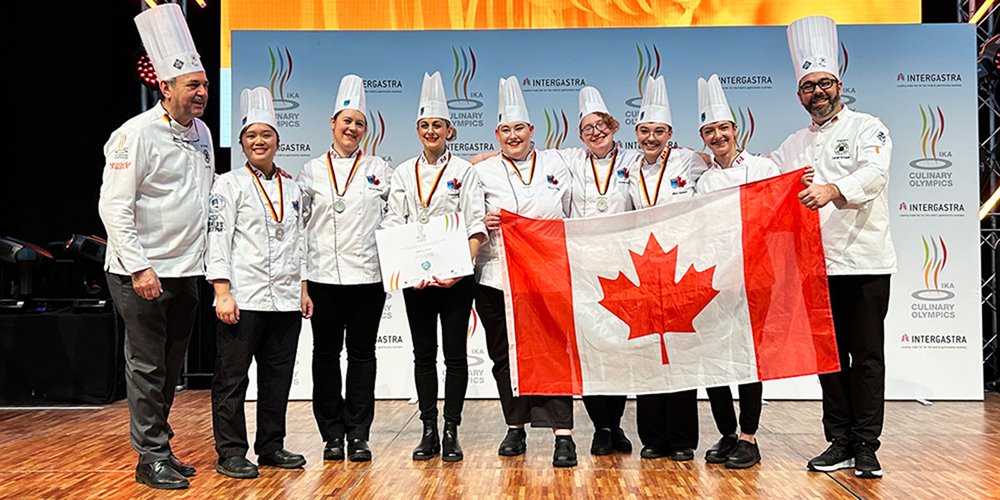 six women in chef's clothes stand on a stage holding a large canadian flag. two male chefs stand on either side of them