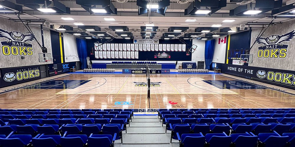 Wide view of an empty indoor gymnasium with a polished court, blue seating, and “OOKS” branding on the walls.