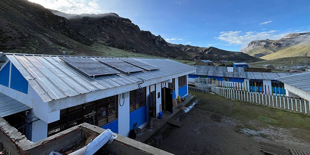 A cluster of blue and white buildings with metal roofs equipped with solar panels is situated in a mountainous area under a clear blue sky, with rugged hills and grassy terrain in the background.
