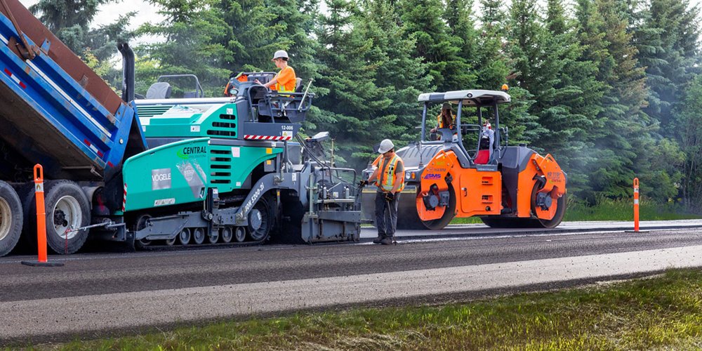 Workers operate heavy machinery to lay and compact asphalt on a road surrounded by dense green trees.