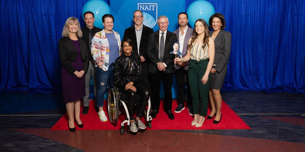 A group of nine people posing for a photo on a red carpet in front of blue curtains and two large blue balloons. A NAIT (Northern Alberta Institute of Technology) logo is visible in the background. One person is seated in a wheelchair, with the others standing around them.