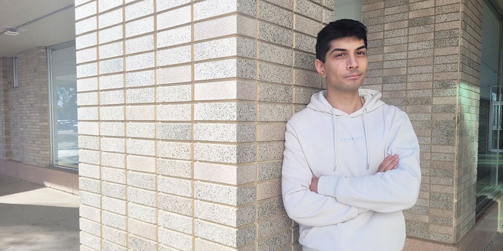 Person wearing a light-colored hoodie stands with arms crossed, leaning against a corner of a brick building with large windows and an outdoor walkway visible.