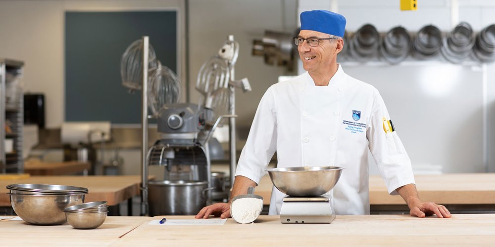 Chef in a white uniform and blue hat stands at a kitchen workstation with metal mixing bowls, a digital scale, and a mound of flour, with commercial baking equipment visible in the background.