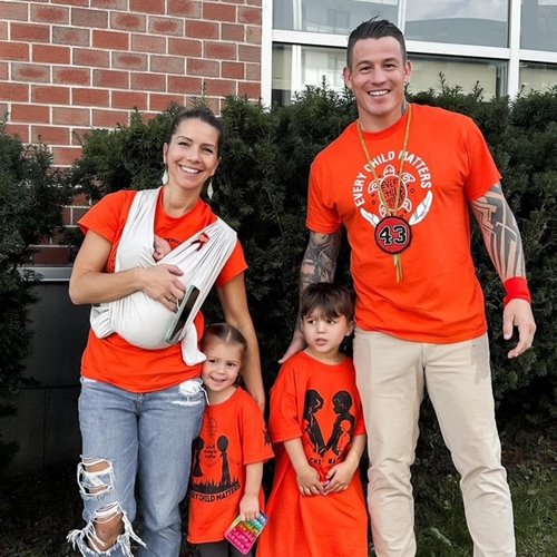 Group of people wearing orange 'Every Child Matters' shirts standing outdoors in front of a brick building, with one person holding a baby and two children standing in front.