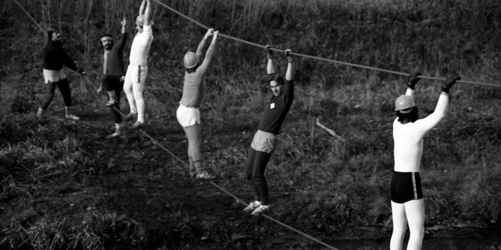 Six individuals crossing a rope bridge outdoors, balancing on a lower rope with their feet and holding an overhead rope with their hands, set in a grassy, uneven terrain.