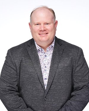 a man in a charcoal coloured blazer and blue and white patterned shirt smiles for a photo