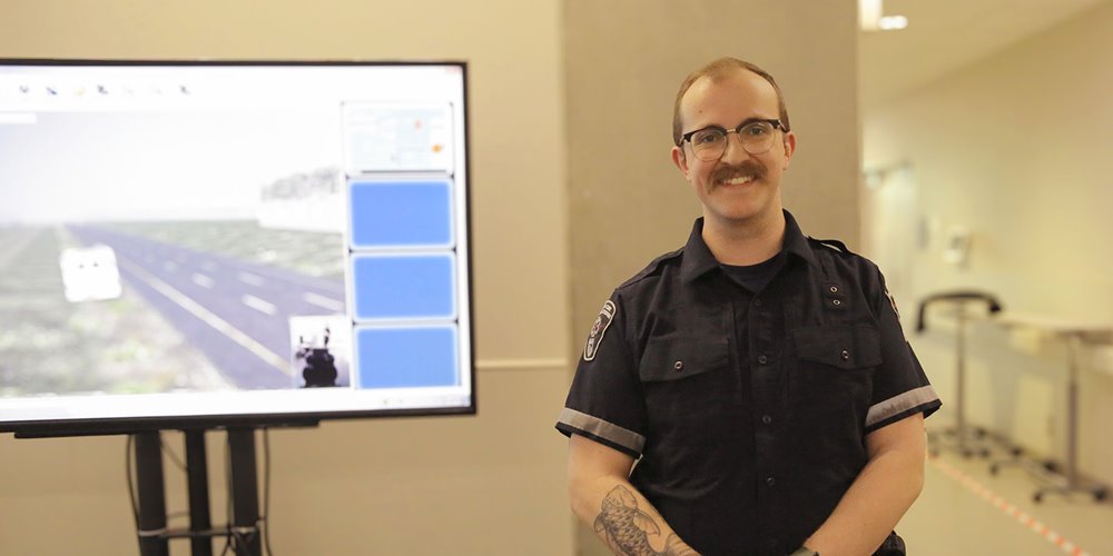 a man in a paramedic uniform stands next to a large monitor in a health care facility smiling and posing for a photo