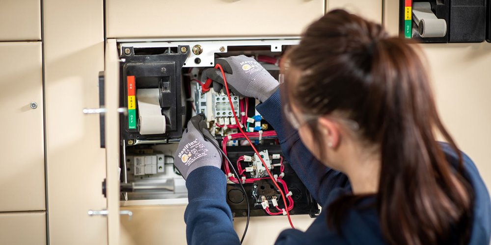 An electrician wearing gloves works on an electrical panel, handling wires and components with tools amid switches and circuit breakers.
