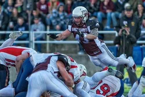 Saint Mary's football player wearing jersey number 43 leaps through the air during a game against a team in white uniforms with red and blue accents, with spectators watching in the background.