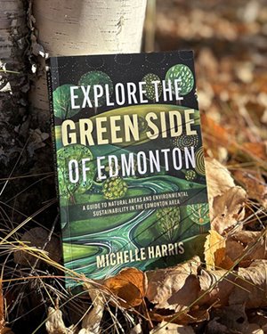 A book titled “Explore the Green Side of Edmonton” by Michelle Harris rests against a tree trunk on a bed of fallen leaves. The cover features green and black artwork with stylized trees and a winding road, and includes the subtitle: “A guide to natural areas and environmental sustainability in the Edmonton area.”