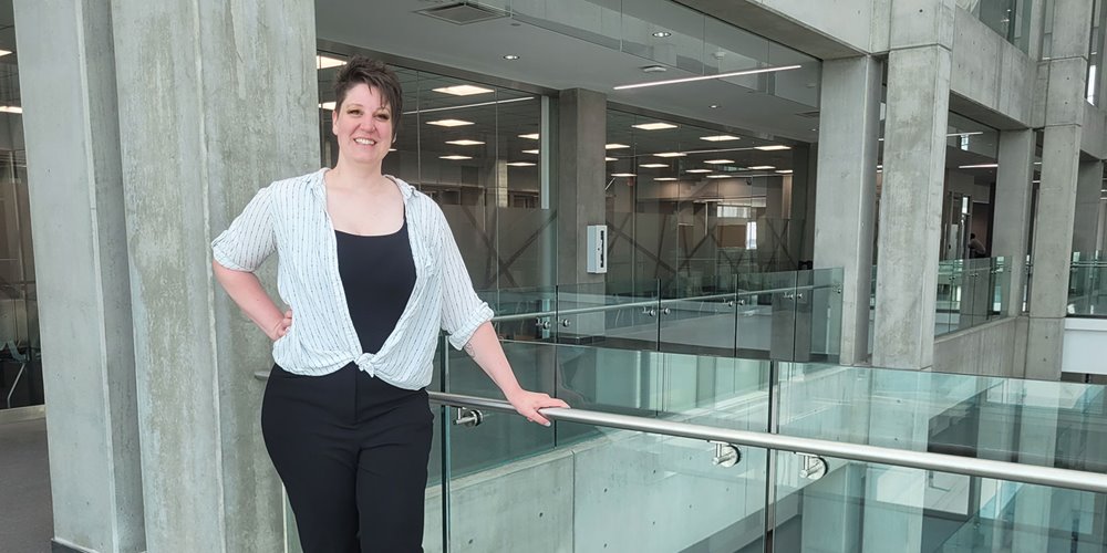 a woman in a white blouse and black shirt leans on a glass railing in a modern looking building and smiles for a photo