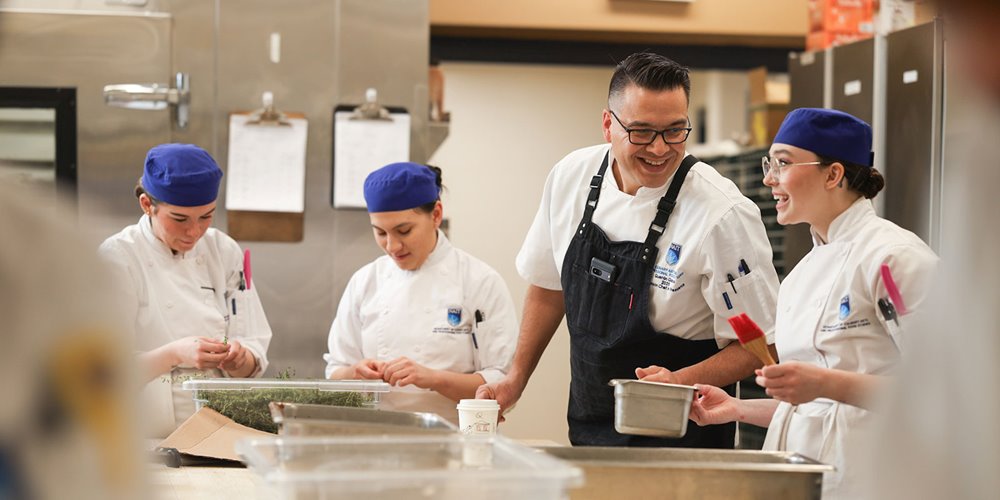 A group of culinary students in white chef uniforms and blue hats are gathered around a stainless steel counter, engaging with an instructor wearing a black apron in a professional kitchen setting.