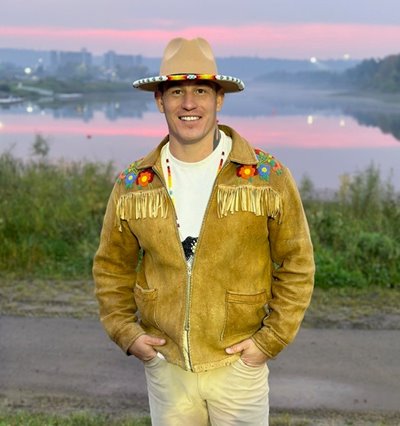 Person wearing a tan suede fringe jacket with floral embroidery and a beige hat stands near a body of water at sunset, with a cityscape in the background under a pink and purple sky.