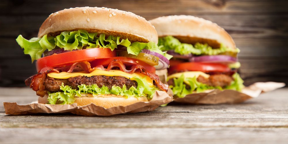 Two hamburgers on a wooden surface, each with sesame seed buns, lettuce, tomato slices, cheese, bacon, and beef patties, partially wrapped in brown paper.