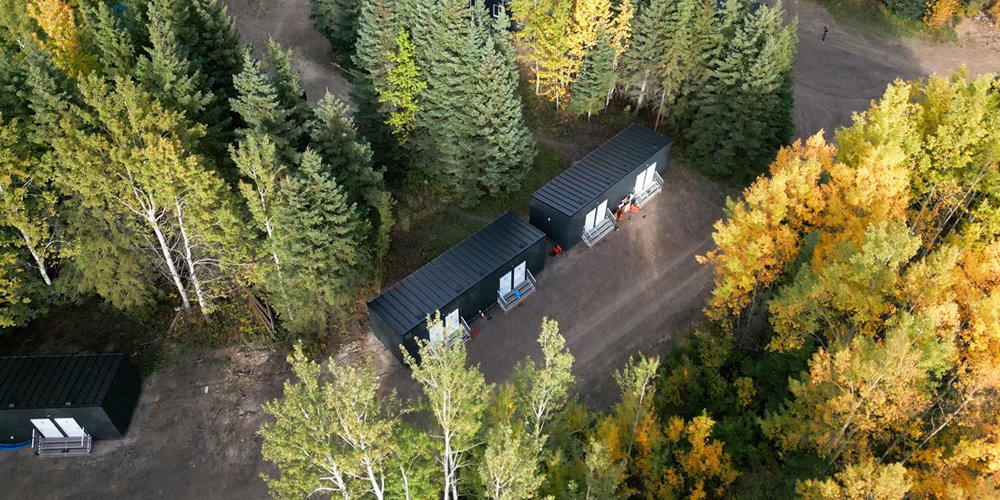 An aerial view of a forest with a small group of black shelters made from shipping containers. The trees are a mix of green and yellow, indicating autumn.