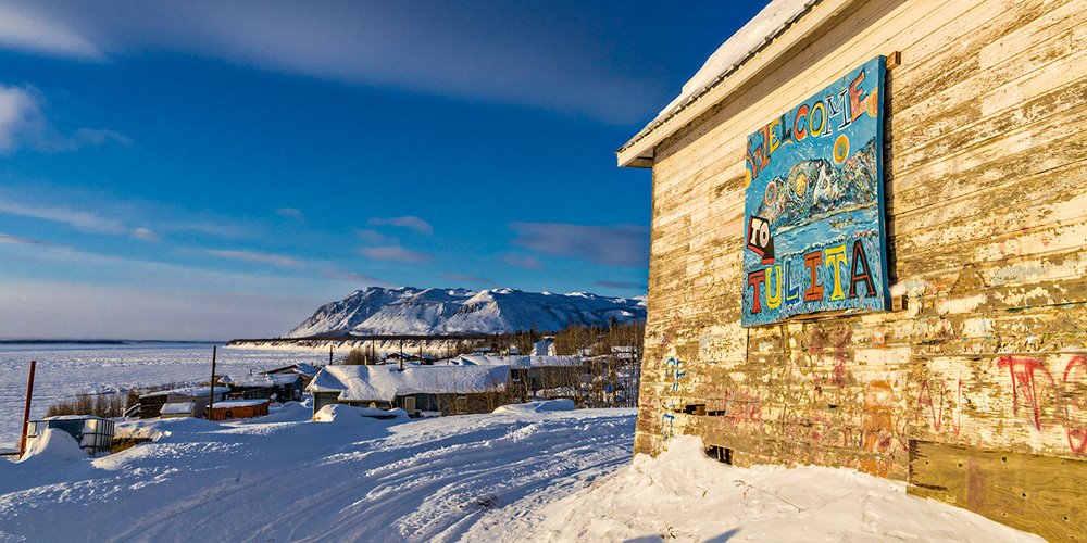 Snow-covered riverside community scene with a weathered building displaying a colorful “Welcome to Tulita” sign and mountains in the background.