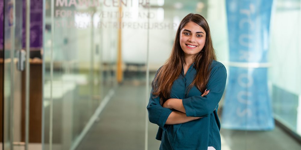 a woman in a blue long-sleeve shirt crosses her arms and smiles as she poses for a photo in a modern-looking building featuring glass walls