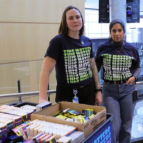 Two individuals wearing matching navy blue T-shirts with the repeated phrase “THIS MATTERS” in green and white lettering stand beside a cart filled with boxes of assorted packaged items. The setting is an indoor public space with glass railings and modern architecture in the background.