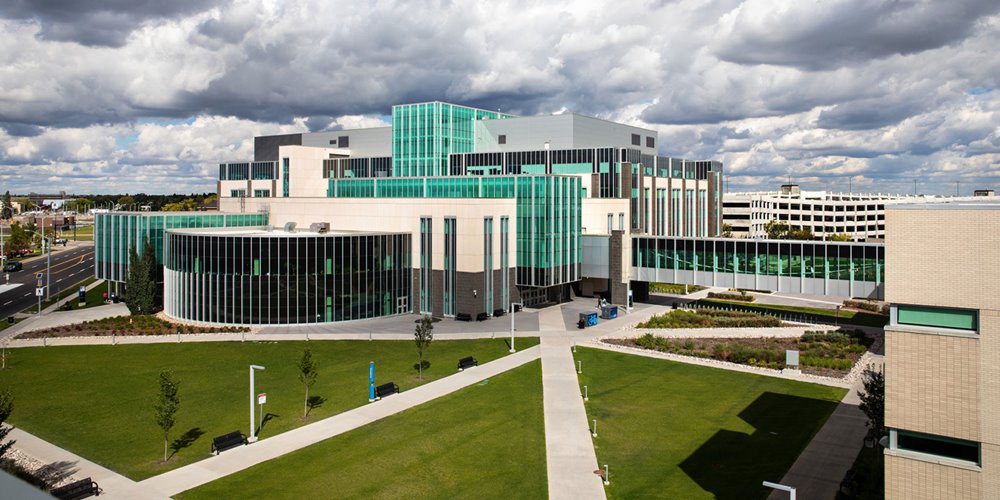 A modern multi-story building with large green-tinted glass panels and beige exterior walls is surrounded by landscaped green lawns, pathways, and trees under a partly cloudy sky.