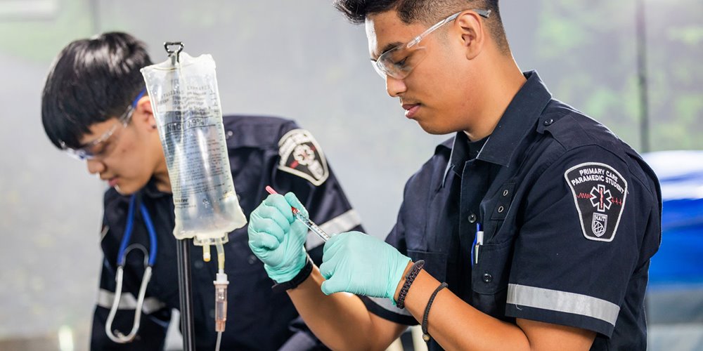 Two paramedic students in uniform practicing medical procedures; one prepares a syringe while the other monitors an IV bag, both wearing gloves and protective eyewear.