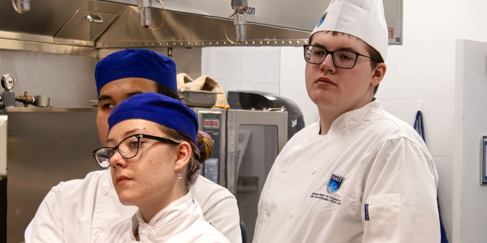 Three individuals in white chef uniforms and hats stand in a kitchen setting with equipment like an oven and a stainless steel hood in the background.