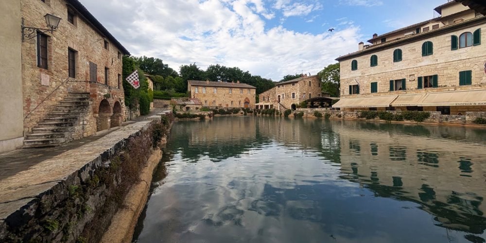 Scenic view of a European village with stone buildings and arched windows surrounding a calm body of water, featuring ivy-covered walls and a staircase leading to the water under a partly cloudy sky.