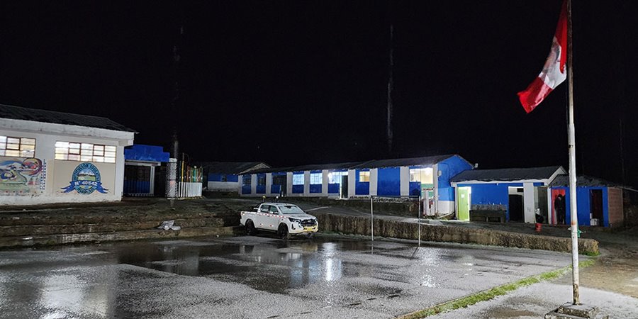 A nighttime photograph showing a series of buildings with blue and white exteriors and lit windows. A white pickup truck is parked on a paved area in front of the buildings. A Peruvian flag on a tall pole is visible in the foreground right, against a dark sky.