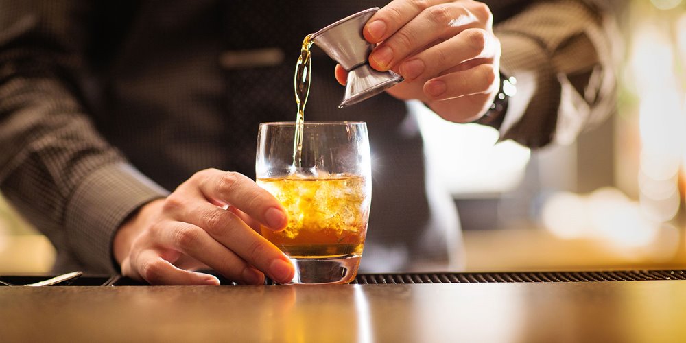 photo of a bartender pouring a dark amber liquid from a measuring cup into a glass filled with ice.