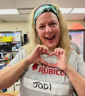 a woman makes a heart with her hands and smiles. she has long hair pulled back with a turquoise bandana and wears a light grey t-shirt