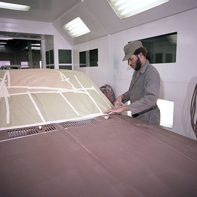 A person is preparing a car for painting inside an automotive spray booth by masking the windshield with tape and protective paper.