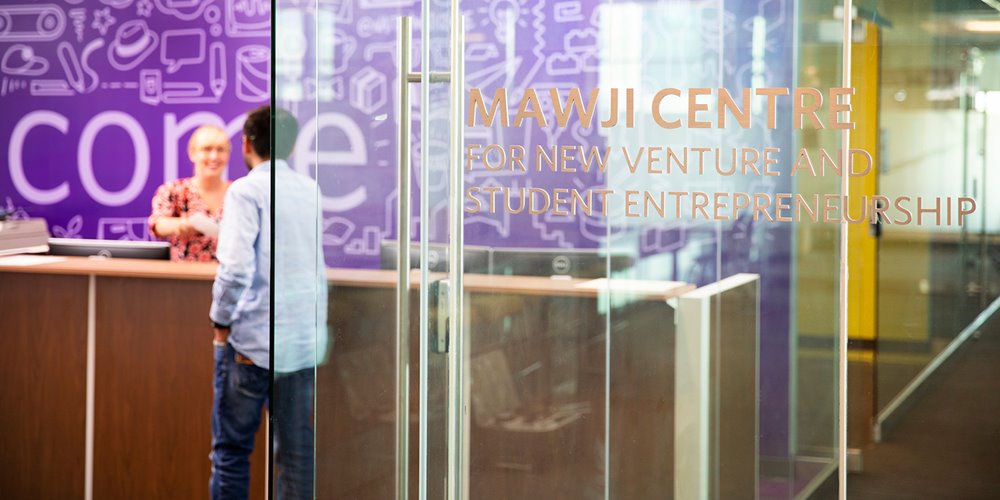 Entrance to the Mawji Centre for New Venture and Student Entrepreneurship, showing a glass door with the centre's name, a reception desk with two people interacting, and a purple wall in the background featuring icons and the word 'connect'.